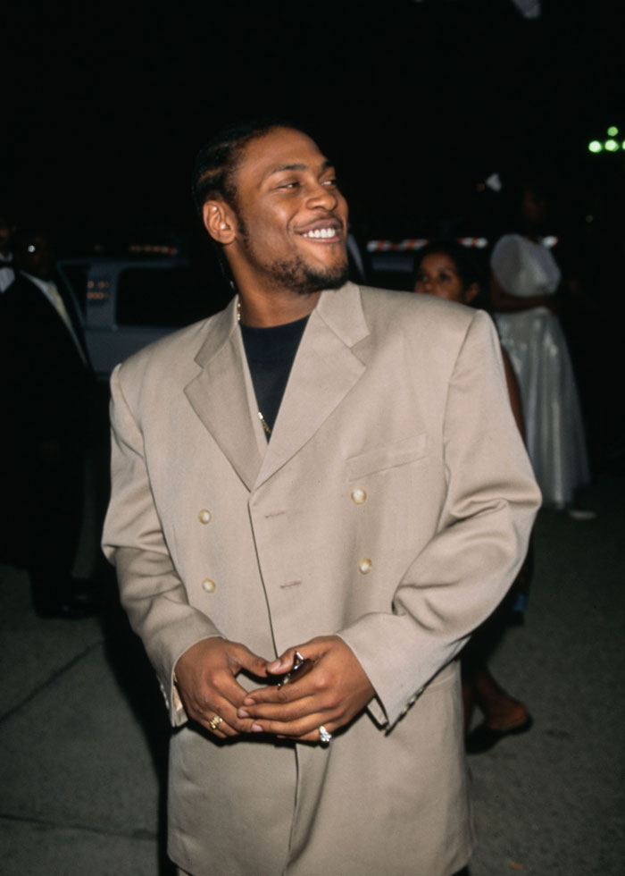 R&B legend D'Angelo smiling in a beige suit jacket at an evening event with people in the background.