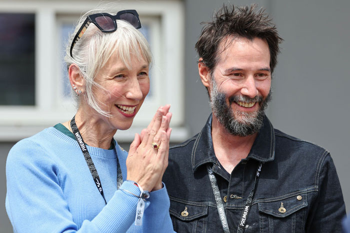 Keanu Reeves smiling with Alexandra Grant, both wearing lanyards, appearing happy and engaged in conversation outdoors.