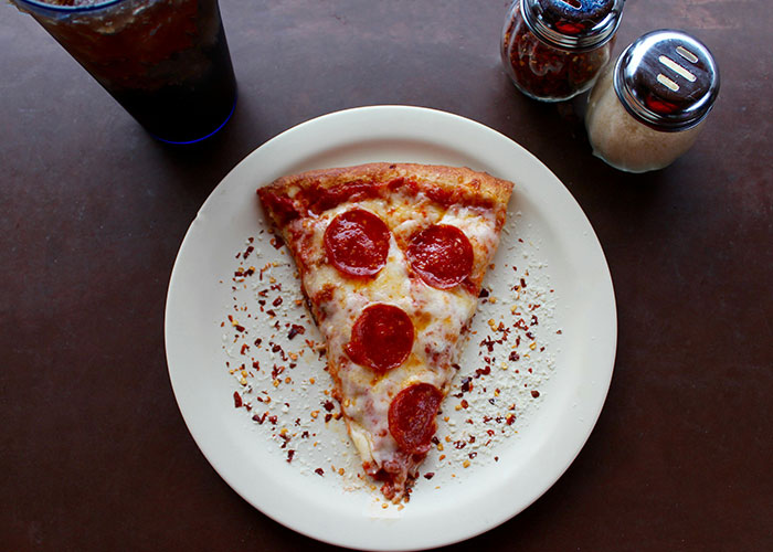 A slice of pepperoni pizza on a white plate with red pepper flakes, salt, and a drink, illustrating loopholes against horrible bosses.