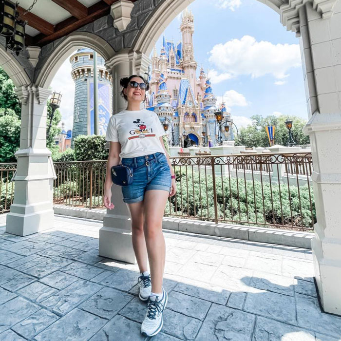 Pregnant Disney superfan posing in sunglasses and shorts near a castle at a Disney theme park on a sunny day.