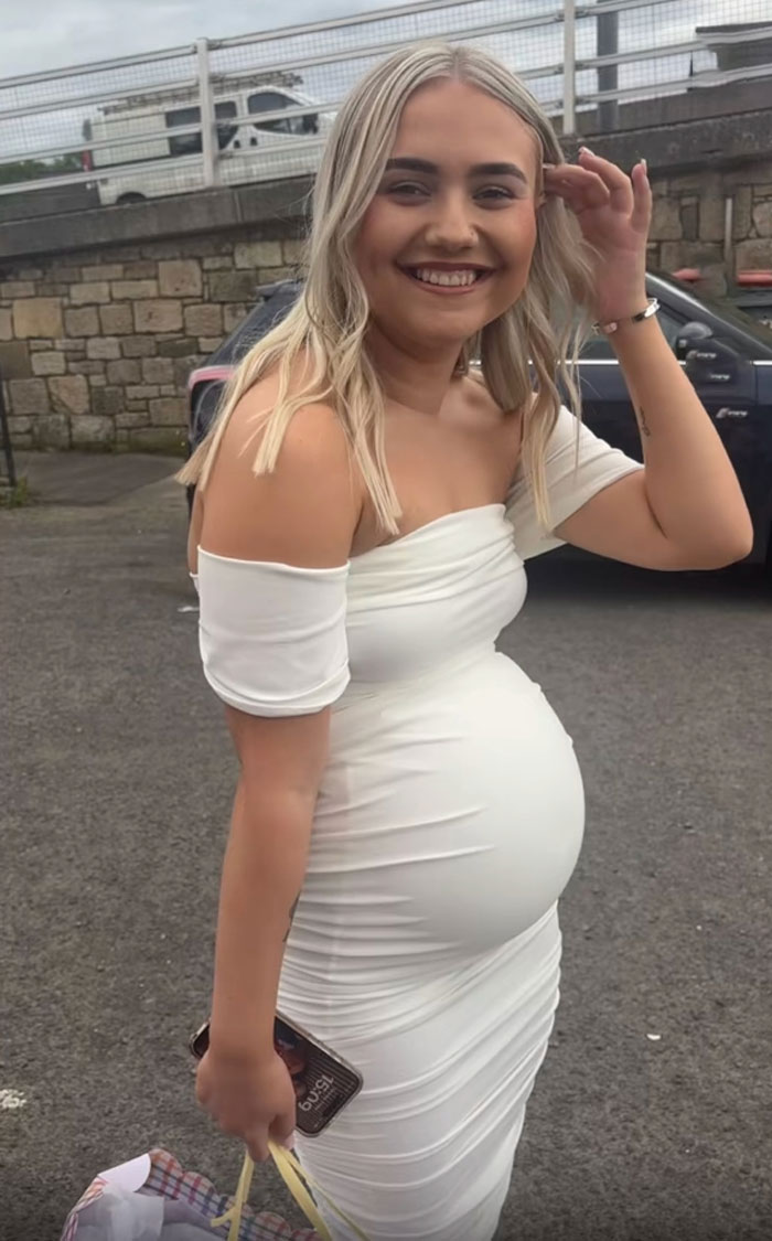 Pregnant woman smiling outdoors in a white dress, linked to outrageous 10-month lie about baby actually being a doll.