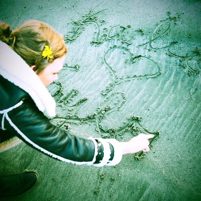 A woman with a yellow flower in her hair writes on sand, symbolizing Taylor Swift lesbian relationship rumors with Karlie Kloss.