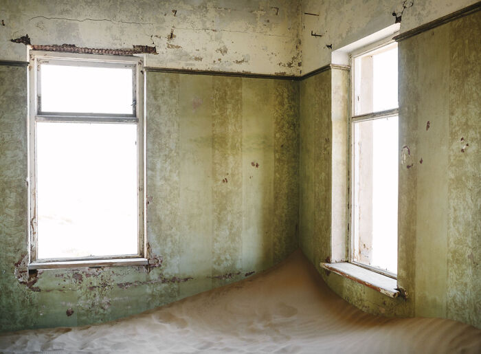 Desert sand slowly filling the corner of an abandoned home with peeling green walls and two large windows.
