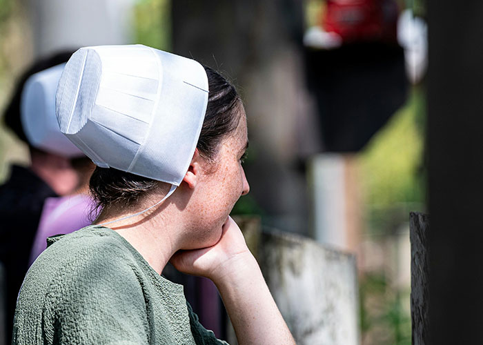 Amish mom in traditional white bonnet and green dress looking away, reflecting on a moment with her child embarrassment.