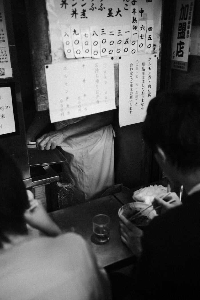 Black and white street photography of people eating at a small restaurant capturing life’s unexpected moments in an urban setting.