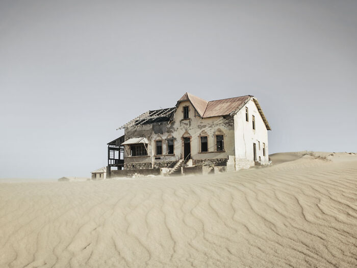 Abandoned home partially buried in desert sand dunes under a pale sky, illustrating sand castles and desert sand invasion.