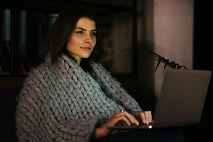Woman wrapped in a chunky knit blanket focused on laptop screen, illustrating intense stalking behavior concept.