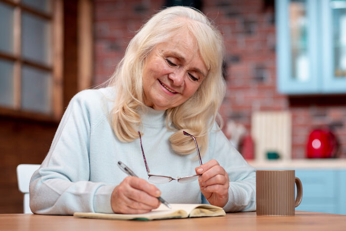 Older woman smiling, holding glasses and pen, writing in a notebook.