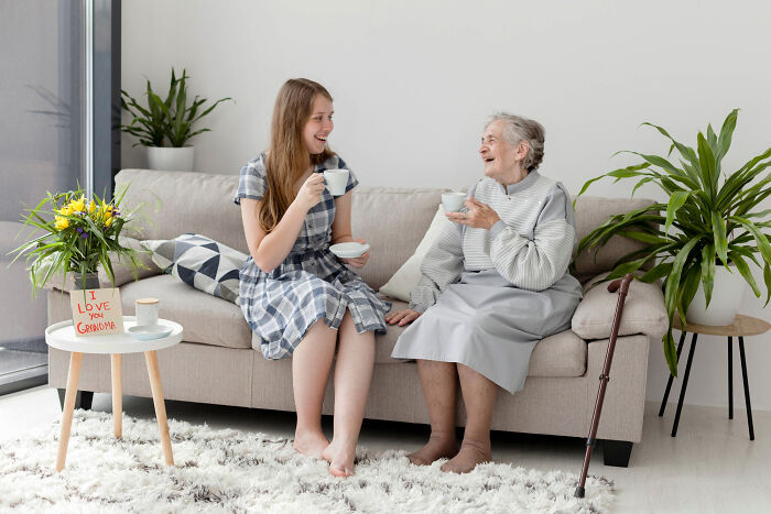 Young woman and elderly patient with dementia sharing a lighthearted moment while drinking tea on a sofa at home.