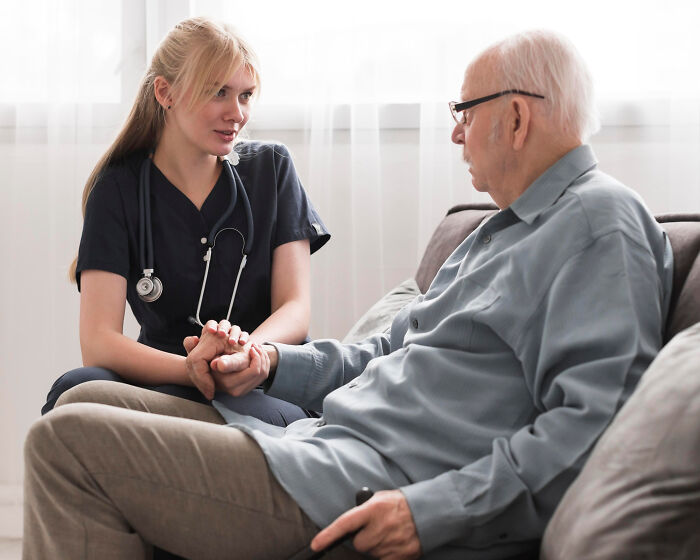 Young female nurse holding hands and talking with elderly dementia patient at home, showing compassionate care and support.