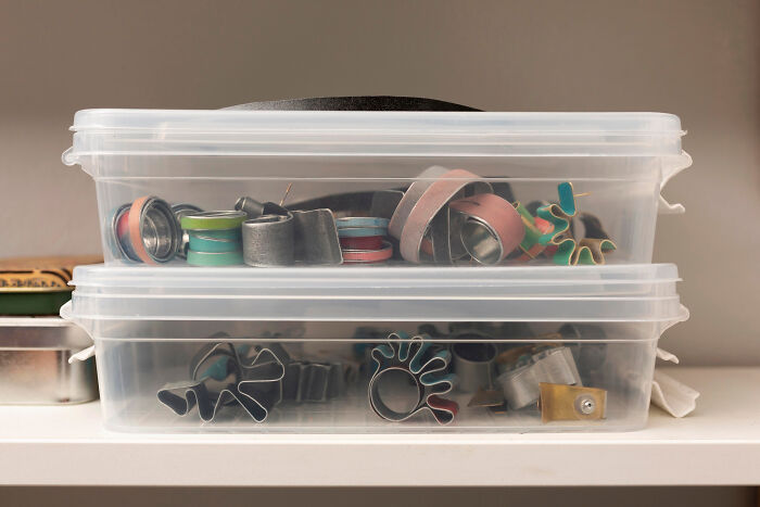 Two clear plastic boxes filled with various metal cookie cutters, stacked on a white shelf for organized storage.