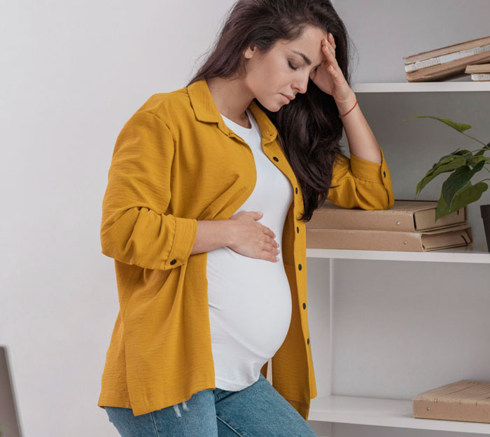 Pregnant woman in a yellow shirt looking tired and sore, holding her belly and forehead in a home setting.