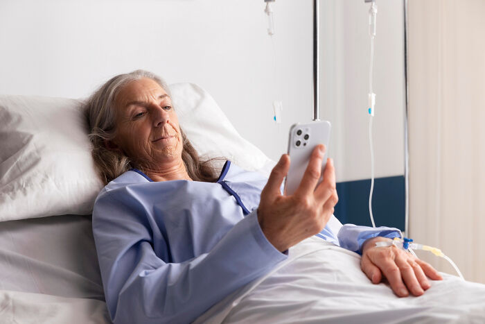 Elderly woman in hospital bed using phone, surrounded by IV drips, depicting scary ghost stories hospital work atmosphere.
