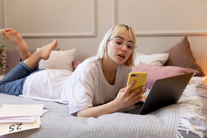 Young woman lying on bed, focused on her phone and laptop, illustrating unhinged stalking behavior stories.