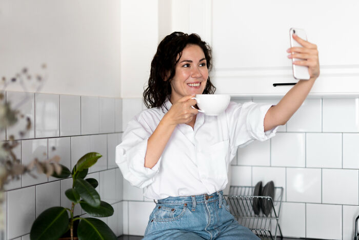 Woman sitting on kitchen counter holding a cup and taking a selfie, illustrating unhinged stalking behavior.