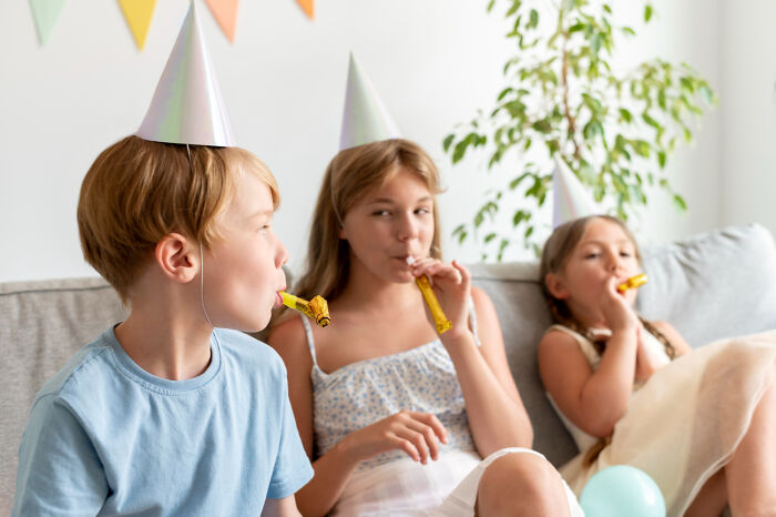 Three children wearing party hats sitting on a couch, celebrating with blowouts, illustrating a painful table for two moment