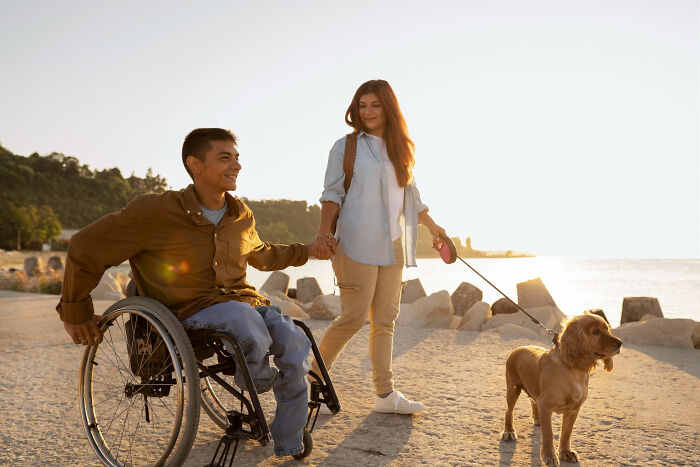 Young man in a wheelchair holding hands with a woman walking a dog, highlighting people with disabilities outdoors.