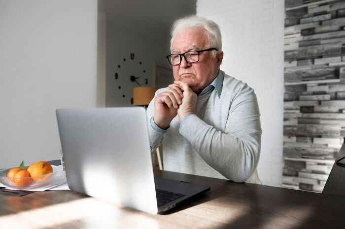 Elderly man with glasses looking puzzled at a laptop.