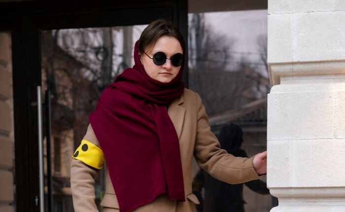 Woman wearing a beige coat, maroon scarf, and sunglasses, appearing awkward while misreading social cues outdoors.
