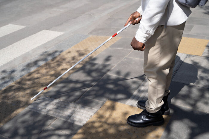 Person with disability using a white cane navigating a sidewalk with tactile paving on a sunny day.