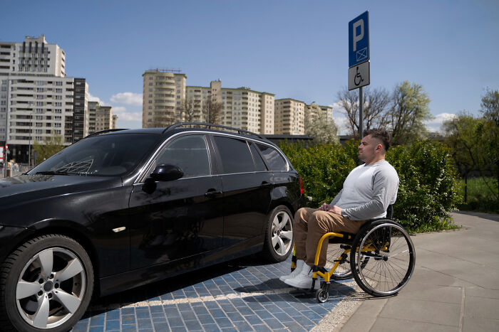 Man in wheelchair next to a car parked in a disabled parking space, highlighting challenges faced by people with disabilities.