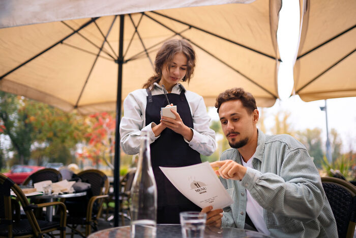 Waitress taking order from customer outside restaurant under umbrella during daytime, highlighting restaurant staff stories.