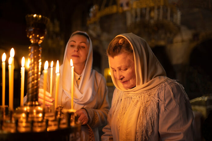 Two women in headscarves lighting candles inside a dimly lit church, capturing a peaceful moment of reflection.