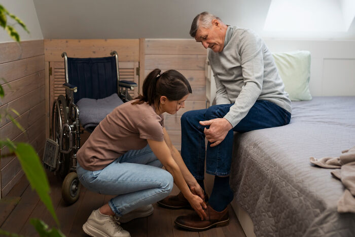 Young woman helping elderly man with disabilities put on shoes near wheelchair in bedroom, highlighting ableist challenges endured.
