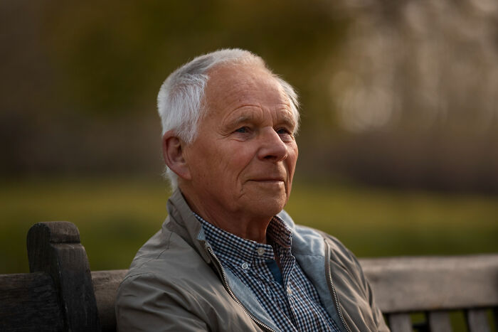 Elderly man sitting outdoors on a bench, reflecting quietly with a calm expression in a natural setting.