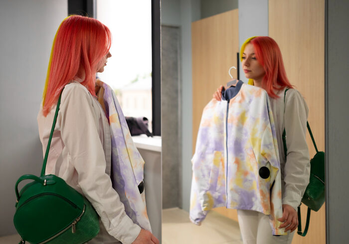Woman with red hair holding a jacket, looking at her reflection in the mirror, illustrating unhinged stalking behavior.