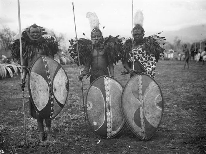 Three warriors in traditional feathered attire with oval shields and spears standing in a field, evoking worst places visited