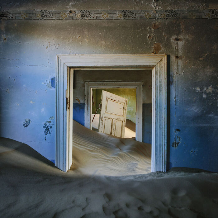 Desert sand filling an abandoned home, covering floors and leaning doors in a surreal sand castle scene.