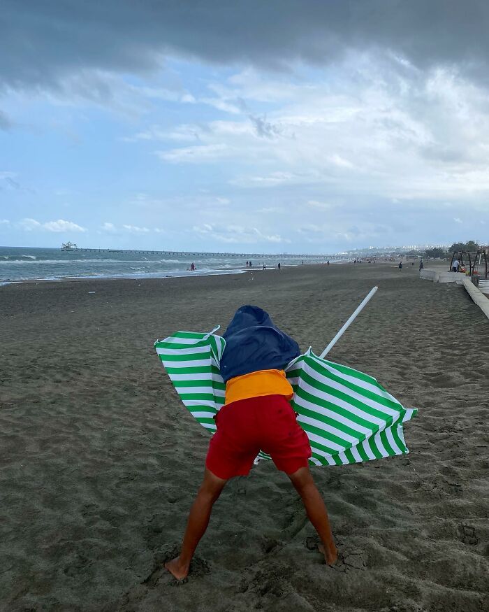 Child on Istanbul's beach struggling with a green and white striped umbrella under cloudy skies, capturing life on Istanbul’s streets.