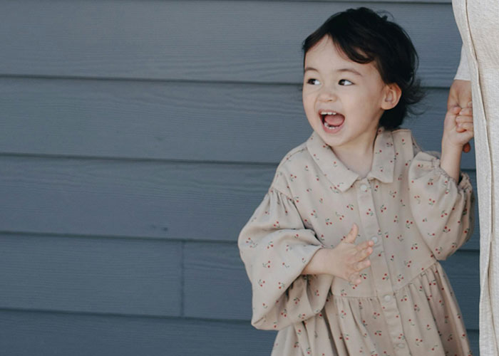 Toddler girl laughing and holding curtain in a dress, illustrating moms share kid embarrassed moments and parenting challenges.