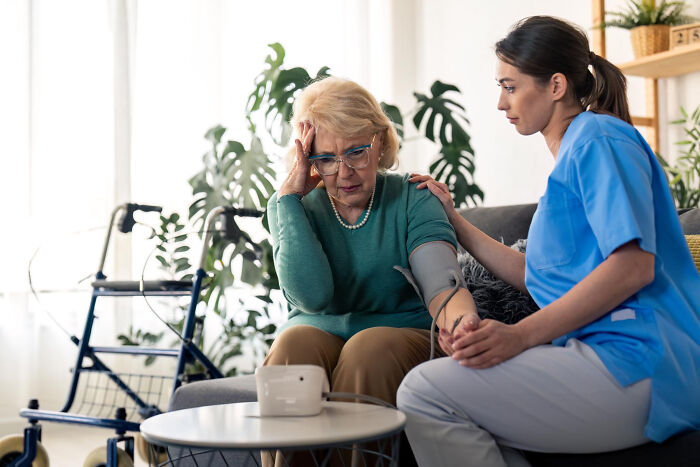 Elderly woman with dementia looking distressed while nurse in blue scrubs offers comfort during health check.
