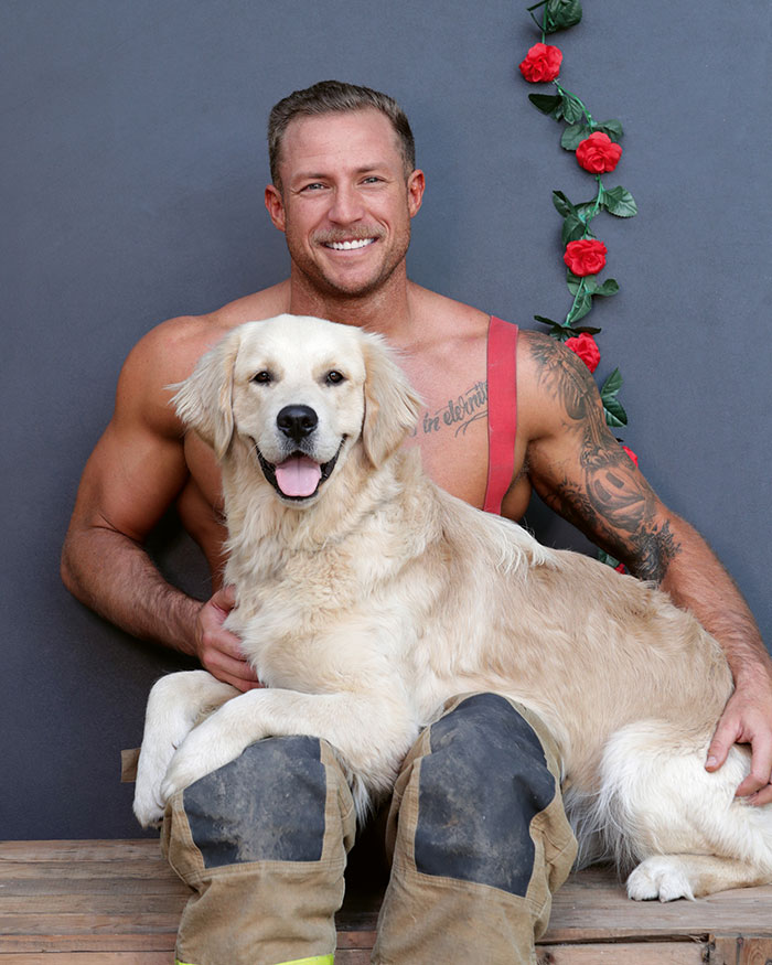 Muscular firefighter wearing suspenders poses with a happy golden retriever dog for Australian calendar photos.