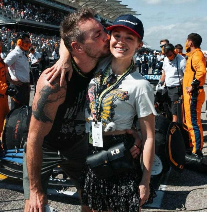 Kristen Bell at an outdoor event, smiling and wearing a cap with "KB" bag during a busy race day scene.