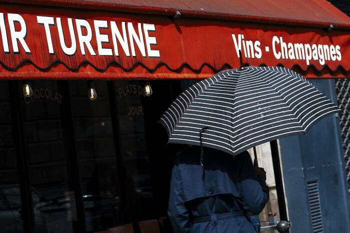 Person holding a striped umbrella walking past a red awning, capturing stunning street photography moments of daily life.