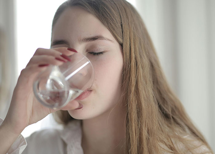 Young woman with long hair using a life hack by drinking water from a clear glass indoors with natural light.