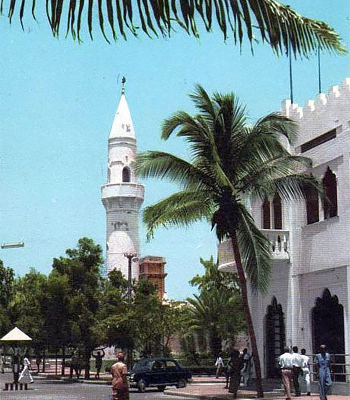 White mosque minaret, palm trees and people on a sunlit street, illustrative of worst places