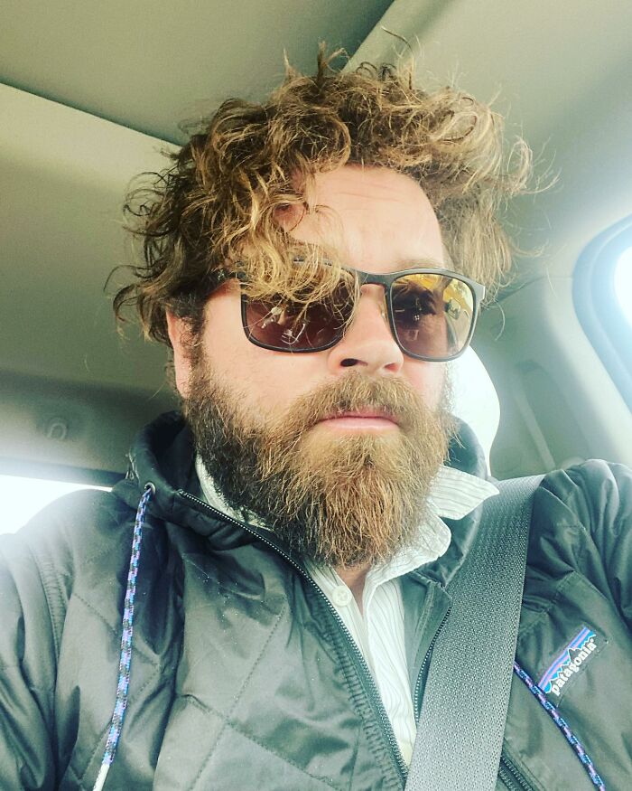 Bearded man with curly hair wearing sunglasses and a dark jacket, sitting inside a car with a seatbelt on.