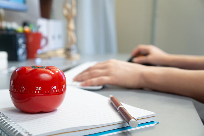 A red tomato-shaped timer on a notebook with a pen, while hands use a computer mouse, illustrating time-saving life hacks.