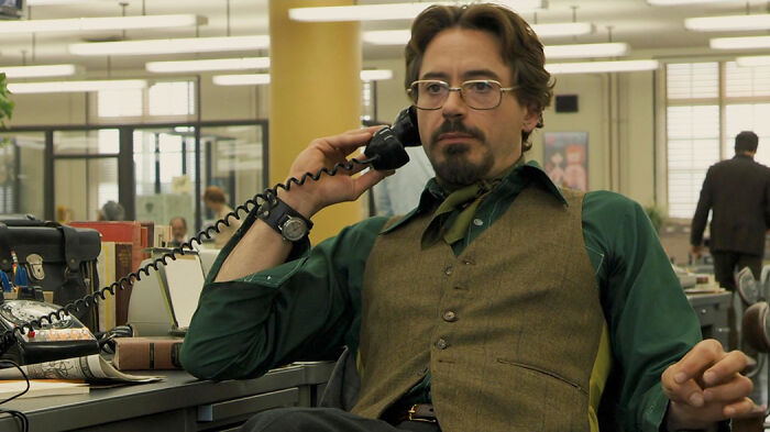 Man in vintage office setting wearing glasses and vest, holding phone, representing movies based on true stories.