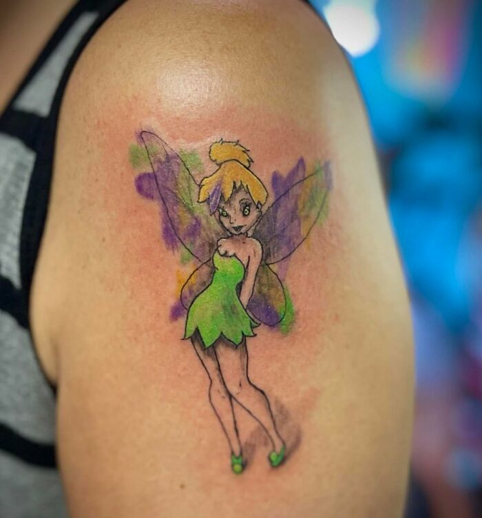 Tattoo of a fairy with purple wings and green dress on upper arm displaying unusual tattoos on bodies.
