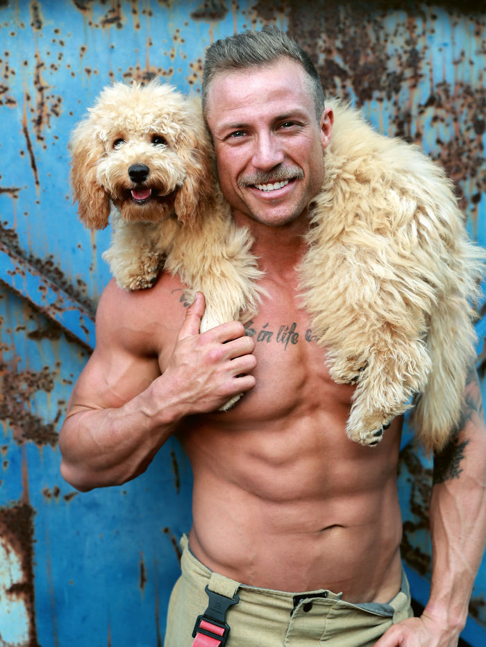 Shirtless muscular firefighter smiling with an adorable fluffy dog draped over his shoulders against a rusty blue backdrop.
