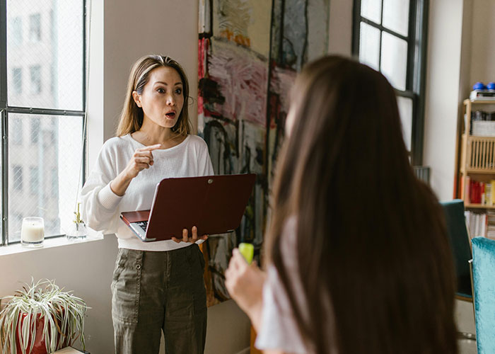 Woman holding a laptop explaining mental health myths to another person in a bright room with large windows.