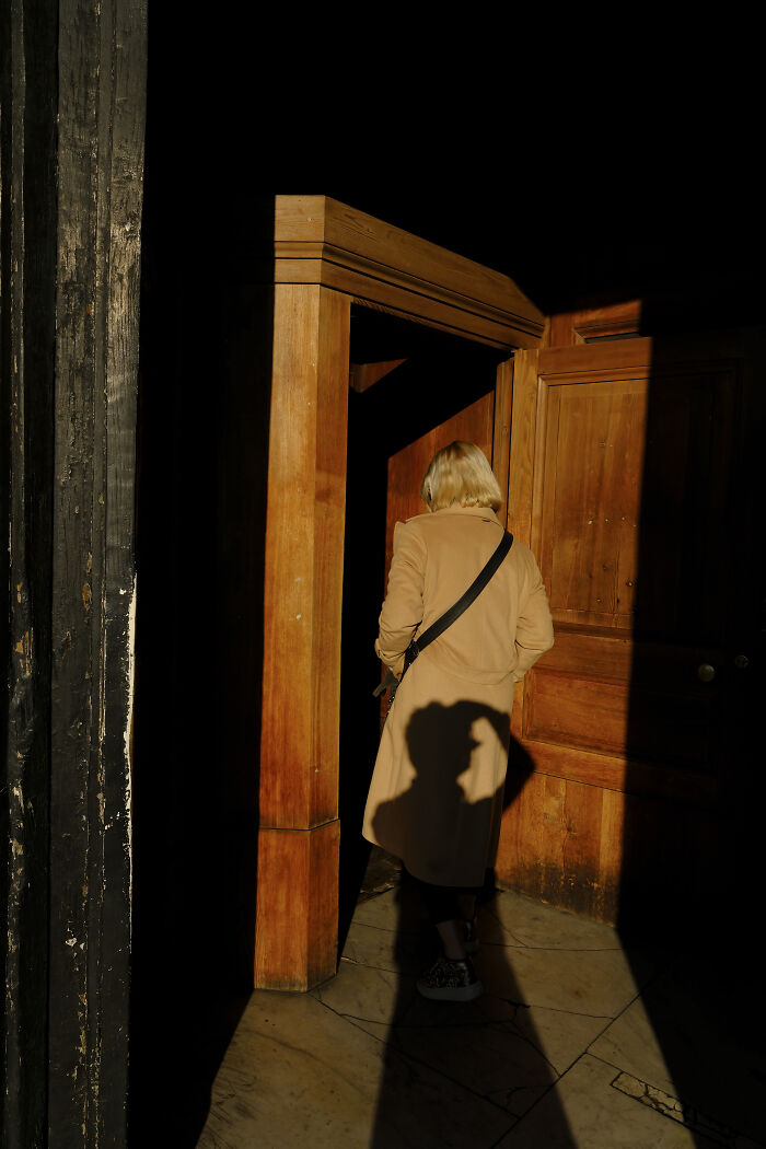 Person in a beige coat entering a wooden doorway with strong shadows in a stunning street photography shot capturing life’s unexpected moments