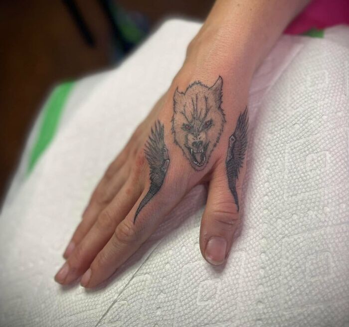 Close-up of a hand tattoo featuring a fierce wolf head and wing designs, showcasing unique tattoos placed on skin.