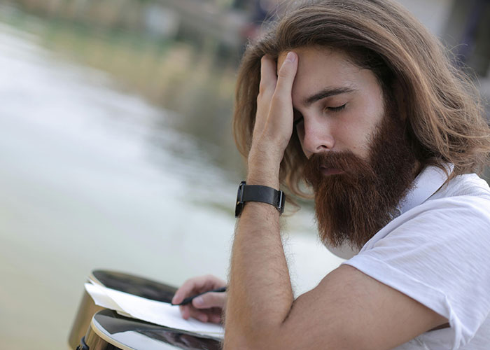 Young man with long hair and beard showing mental distress while writing notes on mental health myths outdoors by water.