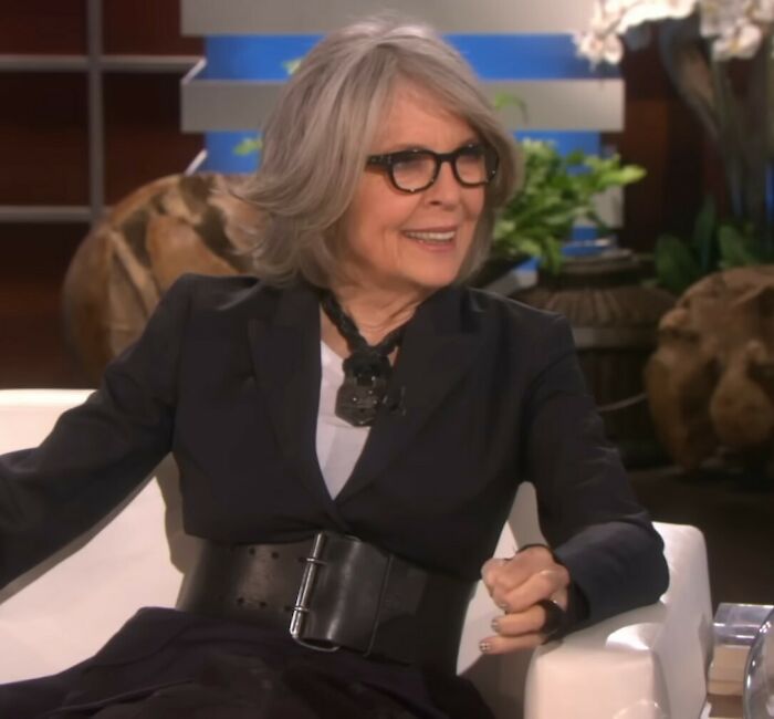 Diane Keaton smiling during a TV interview, wearing black glasses and a black outfit with a wide belt.